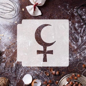 May include: A baking stencil featuring a crescent moon and cross symbol, dusted with flour, rests on a brown surface. The image includes a whisk, a small bag tied with a red ribbon, and scattered hazelnuts, suggesting a baking theme.