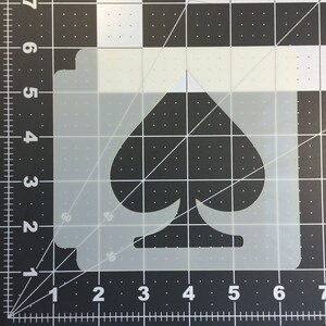 May include: A white plastic stencil of a spade playing card symbol. The stencil is on a black grid background.