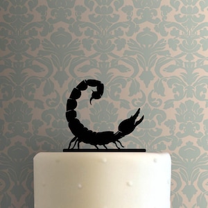 May include: A black silhouette of a scorpion with a curved tail and raised pincers, mounted on a thin horizontal base, positioned atop a smooth white cylindrical surface. The scorpion figure is solid black and features segmented body details.