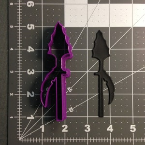 Pode incluir: Cortador de biscoitos de plástico roxo em forma de ponta de flecha com uma pena no lado. O cortador está em um fundo de grade preto e branco.