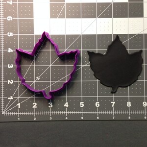 May include: Purple plastic cookie cutter shaped like a maple leaf. The cutter is on a gray cutting mat with white grid lines.