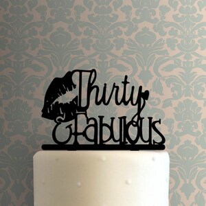 May include: Black cake topper with the words "Thirty & Fabulous" and a lip silhouette. The topper is placed on top of a white cake. The background features a light blue and beige floral pattern.