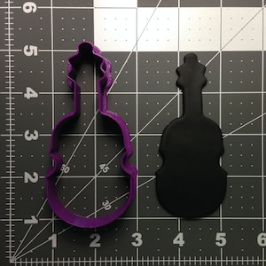Pode incluir: Cortador de biscoitos de plástico roxo em forma de violino. O cortador está sobre um fundo de grade preto e branco. Um recorte de fondant preto em forma de violino está à direita do cortador.