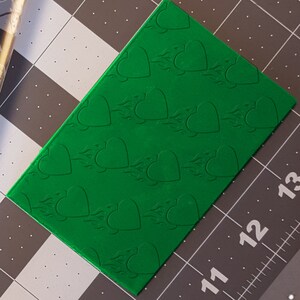 Puede incluir: Una lámina de arcilla polimérica verde con un patrón repetido de corazones en relieve con llamas.
