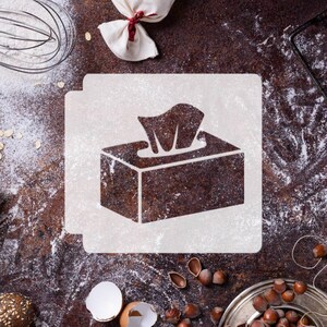 May include: A square stencil featuring a cutout design of a rectangular tissue box with a single tissue emerging from the top. The stencil is white and displays a clear, bold silhouette of the tissue box against a dark, textured background.