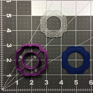 May include: Three cookie cutters in different sizes and colours. The largest cutter is purple and has a scalloped edge. The medium cutter is clear and has a more intricate design. The smallest cutter is blue and has a simple, scalloped edge.