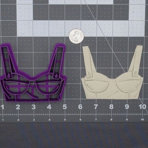 May include: Purple plastic cookie cutter in the shape of a bra. The cookie cutter is on a gray grid surface. Next to it is a beige plastic cookie cutter in the same shape.