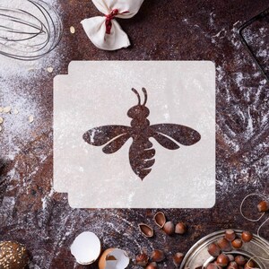 May include: A square white stencil featuring a centered cutout of a bee silhouette. The stencil is positioned on a dark, textured surface dusted with white powder, surrounded by scattered hazelnuts, a metal whisk, and a white cloth bundle.
