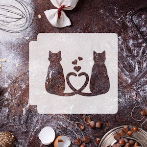 May include: White stencil featuring two cats sitting back to back, their tails forming a heart shape. Small hearts are depicted above the cats. The stencil is placed on a brown surface with flour scattered around it.
