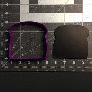 May include: A purple plastic cookie cutter shaped like a slice of bread. The cutter is on a black cutting mat with white grid lines.