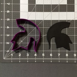 Puede incluir: Un cortador de galletas de plástico morado con forma de casco espartano. Un casco espartano de fondant negro se muestra junto al cortador.