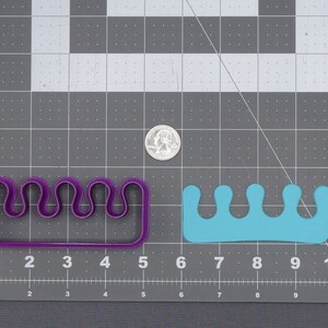 May include: Two cookie cutters, one purple and one blue, shaped like a row of five rounded peaks. The cutters are on a grey grid background.