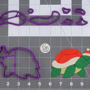 May include: Purple plastic cookie cutters in the shapes of a turtle, a shell, and a Christmas hat. The turtle cookie cutter has a green shell and a red hat.
