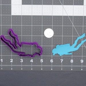 May include: Two plastic cookie cutters in the shape of a scuba diver. The cutter on the left is purple and the cutter on the right is light blue.