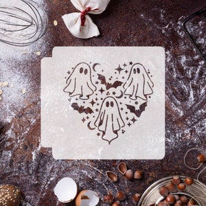 May include: A white stencil with a heart-shaped design featuring three ghosts, bats, stars, and crescent moons. The stencil is on a brown surface with flour scattered around it.