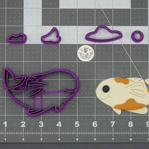 May include: A collection of purple cookie cutters in various shapes, including a cloud, a circle, and a fish. A finished cookie in the shape of a fish is also present. The fish cookie is white with orange and black accents. The image is on a gray grid.