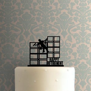 May include: A black cake topper featuring a silhouette of a window washer suspended by ropes against a grid of building windows. The text "HAPPY RETIREMENT" is integrated into the lower right section of the structure, creating a decorative topper.