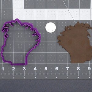 May include: Purple plastic cookie cutter in the shape of the state of Wisconsin. The cutter is on a gray grid background.