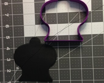 May include: Purple plastic cookie cutter shaped like a vintage gumball machine. The cutter is on a grey grid surface. A black plastic cookie cutter in the same shape is next to it.