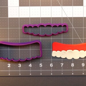 May include: Three cookie cutters, one purple, one red and white, and one white. The purple cutter is shaped like a banner with scalloped edges. The red and white cutter is shaped like a banner with scalloped edges and a white cloud-like design. The white cutter is shaped like a banner with scalloped edges.