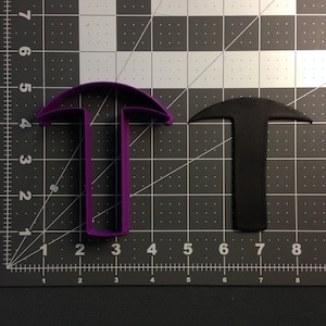 May include: Two plastic cookie cutters in the shape of a hammer. The cutter on the left is purple and the cutter on the right is black.
