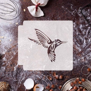 Peut inclure: Un pochoir blanc avec un motif de colibri brun. Le pochoir est sur une surface brune avec de la farine et d'autres ingrédients de cuisson.