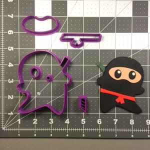 Può includere: Un set di tagliapasta in plastica viola a forma di ninja, nuvola, spada e un piccolo cerchio. I tagliapasta sono su uno sfondo a griglia grigia.