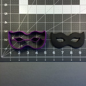 May include: A purple plastic cookie cutter and a black fondant mask, both shaped like a masquerade mask. The cookie cutter is on the left and the mask is on the right.