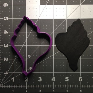 May include: Purple plastic cookie cutter shaped like a seashell. The cutter is on a black and white grid background.
