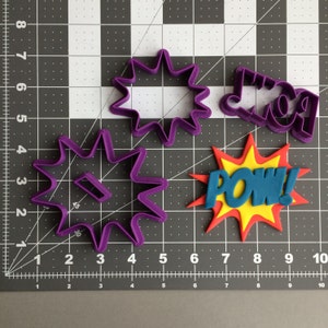 May include: Purple plastic cookie cutters in the shape of a star, a star with a hole in the center, and the word "POW!" in a comic book style. The cutters are on a gray grid background.