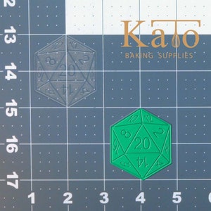May include: A green plastic 20-sided die with numbers 1 through 20 on each face. The die is on a gray grid background.