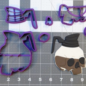 May include: A set of four purple plastic cookie cutters shaped like a skull, a coffee pot, and two coffee beans. The skull is wearing a black bandana and has a brown face.