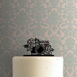 May include: Black silhouette of a camera with floral accents and the name "BONNIE" on a white cake.