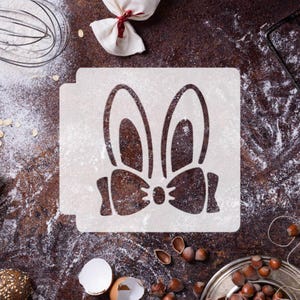 May include: A white stencil featuring a bunny face with large ears and a bow tie, dusted with flour. The stencil is on a dark brown surface with baking ingredients like hazelnuts, an eggshell, and a whisk.