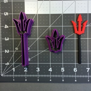 May include: Three cookie cutters in the shape of a trident. The cutters are purple, red, and black. The cutters are on a gray grid background.