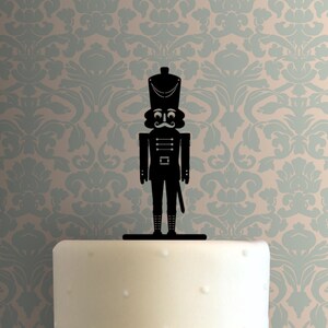 May include: Black silhouette of a nutcracker soldier cake topper. The nutcracker is wearing a tall hat and has a mustache.