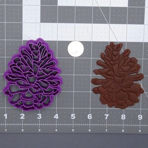 May include: Two pine cone cookie cutters, one purple and one brown, on a gray cutting mat with white grid lines.
