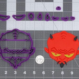 May include: Two purple plastic cookie cutters, one shaped like a devil's face and the other shaped like a human face. The devil cookie cutter is red and has yellow eyes. The cookie cutters are on a gray grid background.