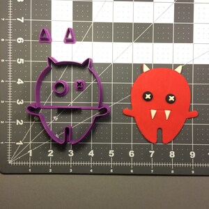 May include: Purple plastic cookie cutter shaped like a cartoon monster with horns, eyes, and teeth. A red fondant cookie is shown next to the cutter.