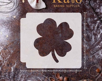 May include: White plastic stencil of a four-leaf clover, with a ruler in inches along the bottom and side of the stencil.  The stencil is on a brown, rustic surface with flour and nuts.