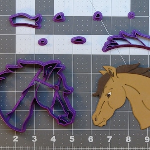 May include: Purple plastic horse head cookie cutter with a brown horse head cutout. The cookie cutter is in the shape of a horse's head with a bridle and mane. The cutout is a brown horse head with a mane and a white eye.