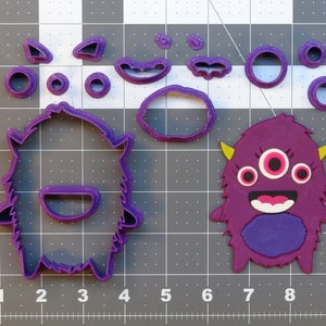 May include: Purple cookie cutters in the shape of a monster with three eyes and a smiling mouth. The set includes a large monster cutter, a smaller monster cutter, and several smaller cutters for the eyes, mouth, and horns.