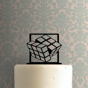 Op de afbeelding: Zwarte silhouet van een Rubik's Cube-taarttopper. De kubus is in een gekantelde positie, met enkele zichtbare vierkanten. De topper staat op een witte taart.