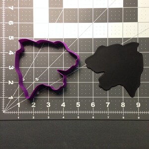 May include: A purple plastic cookie cutter in the shape of a bear, next to a black bear-shaped object. The cutter is on a gray and black grid surface with inch measurements.