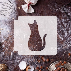 Puede incluir: Una plantilla blanca con la silueta de un gato sentado sobre sus patas traseras. La plantilla está sobre una superficie marrón con harina esparcida alrededor.