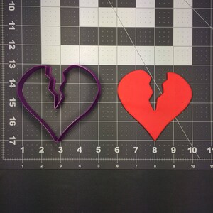 May include: Two broken heart cookie cutters, one purple and one red, on a black and white grid surface.
