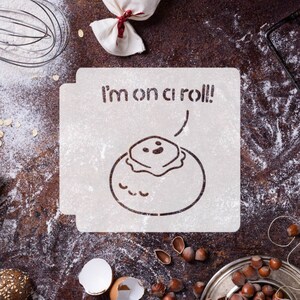 May include: A white stencil with the text "I'm on a roll!" and a cartoon image of a cinnamon roll with a smiling face. The stencil is on a brown surface with flour scattered around it.