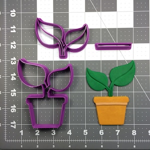 Könnte beinhalten: Zwei violette Plastik-Ausstecher in Form von Topfpflanzen. Ein Ausstecher hat zwei Blätter und der andere vier Blätter. Die Ausstecher befinden sich auf einem grauen Gitterhintergrund.