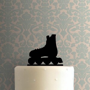 May include: A black roller skate silhouette cake topper. The topper is placed on a white cake with a dotted texture. The background features a light blue and beige floral pattern.