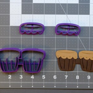 May include: A set of three purple plastic cookie cutters shaped like a pair of bongos and two smaller drums. The bongos are connected and the smaller drums are separate.
